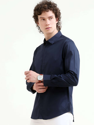 Man wearing a navy blue casual shirt and white trousers, posing and looking to the side in a white studio background.
