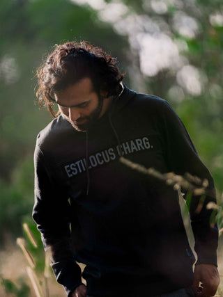 Man wearing a black hoodie with 'ESTILOCUS CHARG.' text, standing outdoors with blurred greenery in the background.