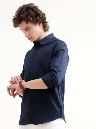 Man wearing a navy blue casual shirt and white trousers, looking at his wristwatch while posing against a white studio background.