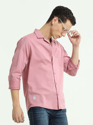 Man wearing a pink casual shirt and blue jeans, adjusting his clear glasses while posing against a white studio background.