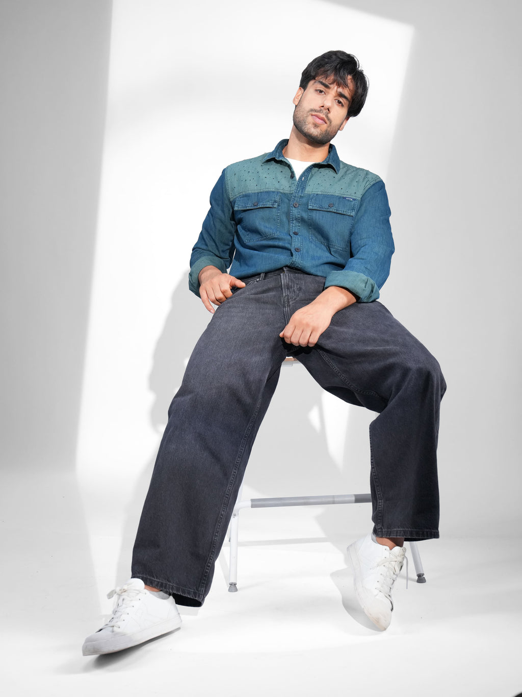 Man sitting on a chair wearing a blue denim shirt and dark jeans against a white background.