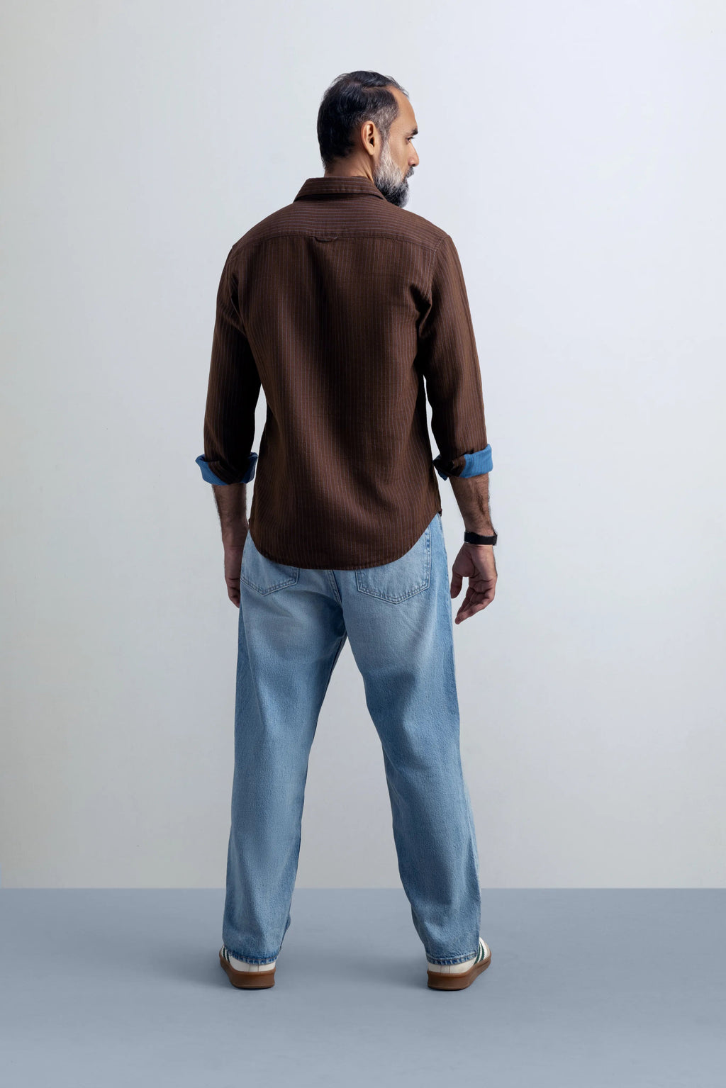 Man wearing a brown shirt and blue jeans standing against a plain background.