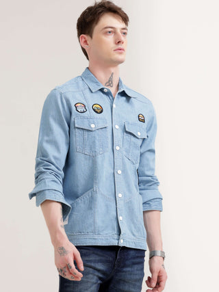 Man wearing a light blue denim shirt with patches on a plain background