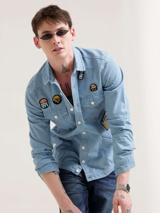 Man wearing a light blue denim shirt with patches on a plain background.