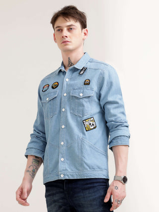 Man wearing a light blue denim shirt with patches on a plain background.