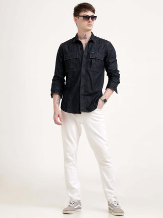 Man wearing a black denim shirt and white pants on a white background.