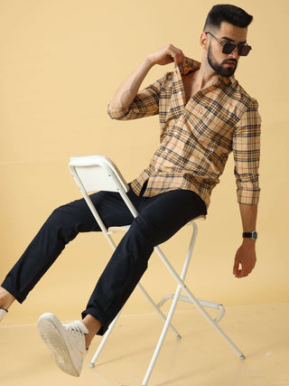 Man sitting on a white chair wearing a checkered shirt, black trousers, and white sneakers, posing in a studio with a light background.