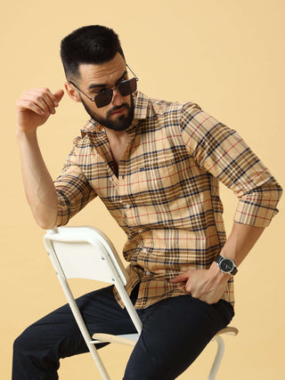 Man wearing a beige checkered shirt sitting on a white chair, posing against a light studio background.