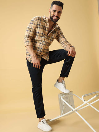 Man wearing a beige checkered shirt and black trousers, standing with one foot on a white chair in a light studio background.